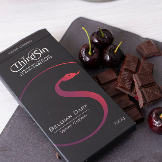Box of Thrice Sin Belgian Dark chocolate with dark chocolate pieces and cherries on a slate board.