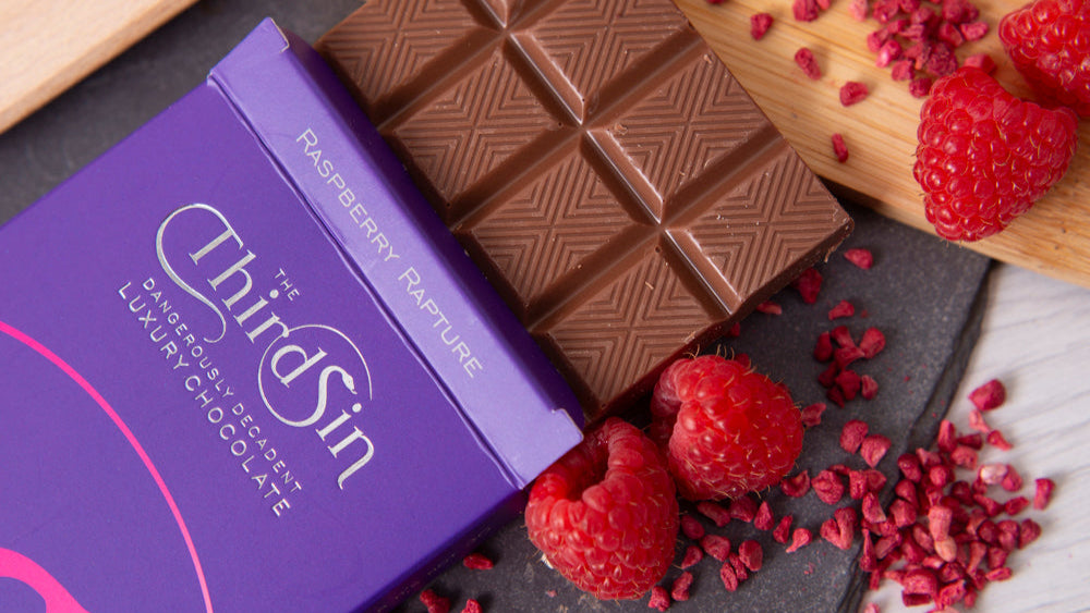 Chocolate bar with raspberries and 'The Third Sin' packaging on a dark surface