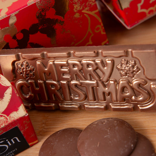 Bronze 'Merry Christmas' chocolate mold on a wooden surface with red and gold decorative elements.