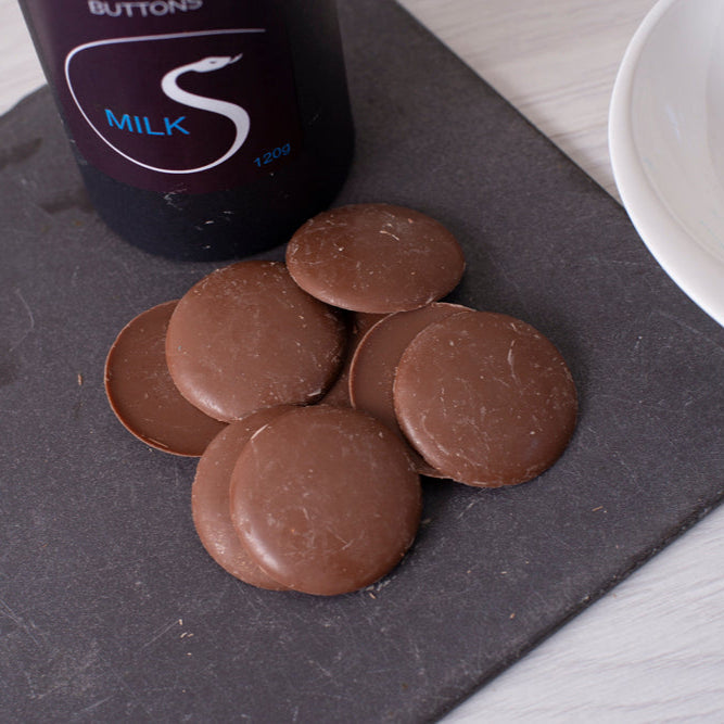 Chocolate buttons on a slate surface with a bottle labeled 'Buttons Milk' in the background.