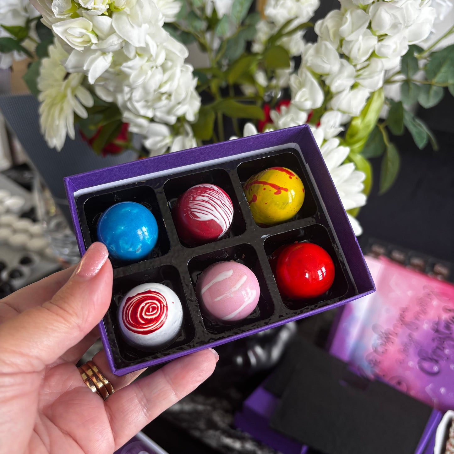 Hand holding a small purple box with colorful spherical treats against a floral background