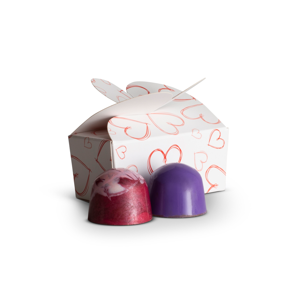 Two purple and pink chocolate truffles in front of a decorative box with heart patterns on a white background.