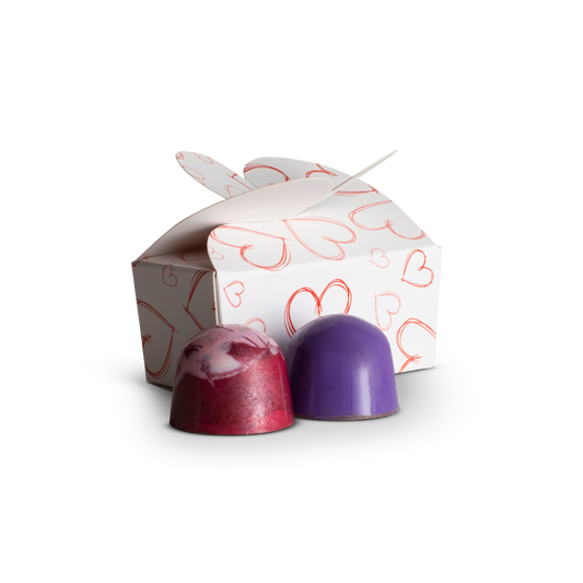 Two purple and pink chocolate truffles in front of a decorative box with heart patterns on a white background.